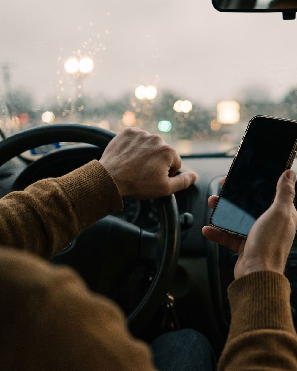 A hand on a steering wheel holding a phone in a parked car at dusk.